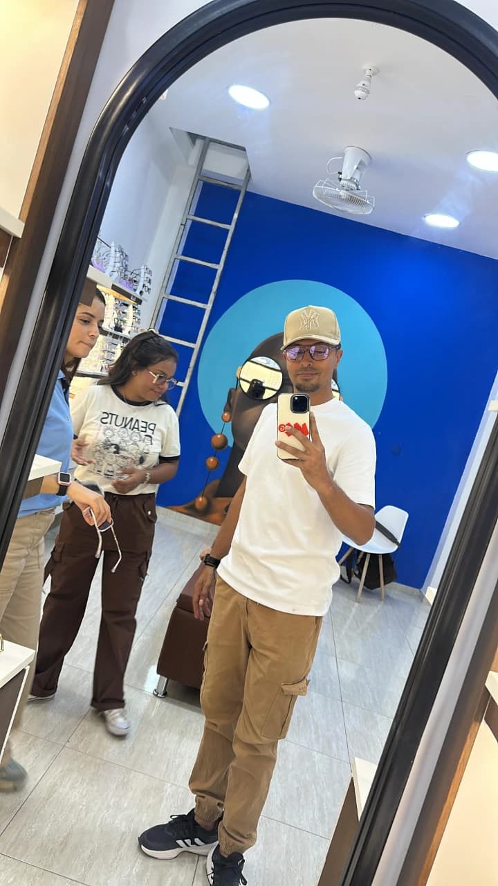 El influencer Maicolteguia visitando Optilux Valledupar en Valledupar. Selfie en el espejo probándose gafas junto al equipo de asesores.